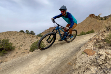 A person riding a mountain bike on a dirt trail through a hilly landscape. The rider is wearing a helmet and colorful attire, leaning into a turn as they navigate the terrain. The background features rocky hills and sparse vegetation under a cloudy sky. 18 Road Trails / North Fruita Desert mountain bike trail.