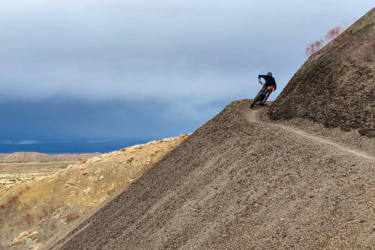 A mountain biker navigates a steep, rugged trail on a rocky hillside, surrounded by expansive desert landscape under a moody sky. 18 Road Trails / North Fruita Desert mountain bike trail.