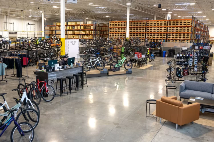 An indoor retail space showcasing a variety of bicycles and cycling gear. The area features rows of bikes displayed on racks, a sales counter with a staff member assisting customers, and clothing displays on hangers. There are also shelves stocked with accessories in the background, and casual seating arrangements, including a couch and chairs, in one corner of the shop. The space is well-lit and has a modern, open feel.