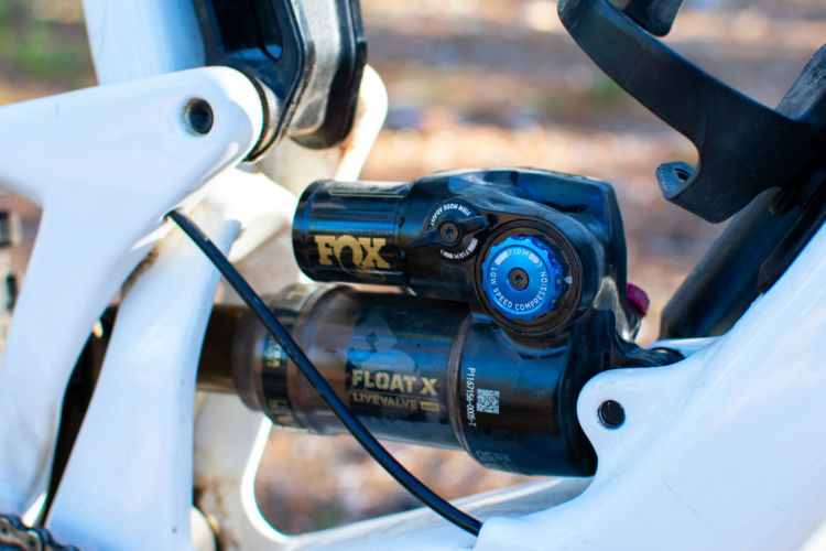 Close-up of a FOX Float X Live Valve shock absorber, featuring adjustable settings for compression and firmness, mounted on a white bicycle frame. The image highlights the intricate design and components of the shock, set against a blurred natural background.