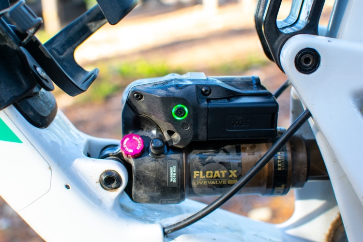 Close-up view of a mountain bike's suspension system, featuring a black shock with an electronic control unit, highlighted by a green indicator light and a pink adjustment knob. The bike frame is white with green accents, positioned in a natural outdoor setting.