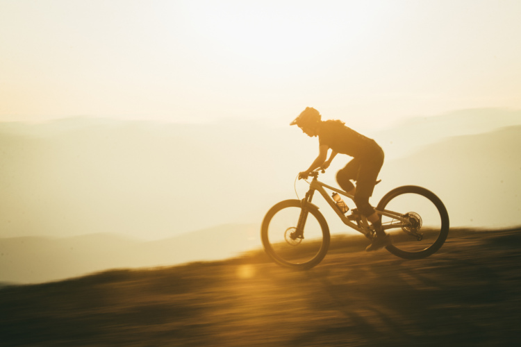 A mountain biker rides along a hillside during sunset, with a warm, golden glow illuminating the scene and soft mountain silhouettes in the background. The biker is in motion, capturing a sense of speed and adventure.