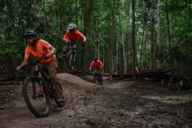 A group of mountain bikers wearing bright orange shirts is captured in a dense forest. One rider is in mid-air performing a jump while two others are on the ground navigating the dirt trail. The scene is surrounded by tall trees and lush greenery, showcasing an active outdoor environment. Legacy Park mountain bike trail.