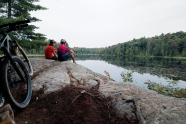 Two people sit on a rocky outcrop by a calm lake, surrounded by lush greenery. One person is wearing a red shirt, and the other is in a purple shirt with a helmet. Mountain bikes are positioned nearby, hinting at a day of outdoor adventure. The scene captures a serene moment of contemplation against a backdrop of natural beauty. Farmer Lake mountain bike trail.