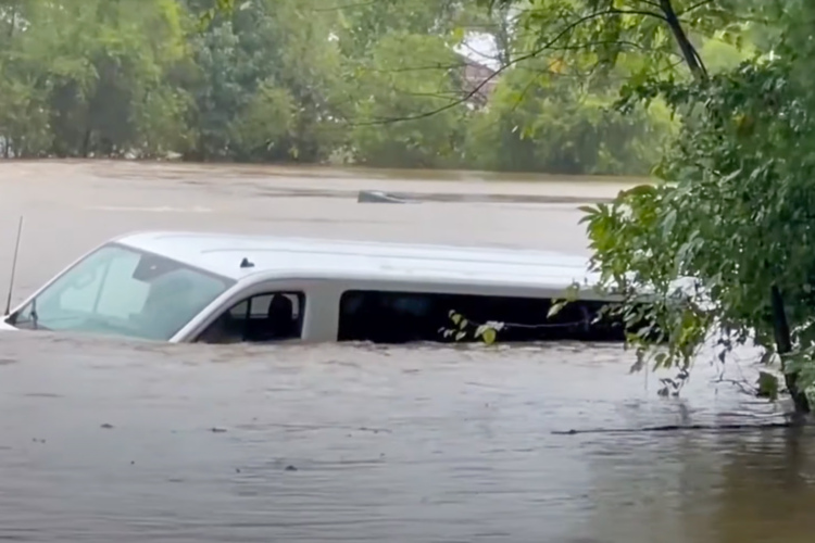 A partially submerged white van surrounded by floodwaters, with trees and a yellow warning sign visible in the background. The scene depicts the effects of heavy rainfall and flooding.