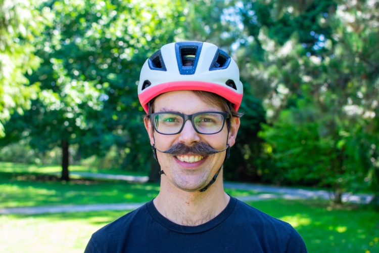 A smiling man wearing glasses and a Specialized Search bike helmet with a pink accent stands outdoors in a park, surrounded by green trees and grass. He has a distinctive mustache and is dressed in a black t-shirt.