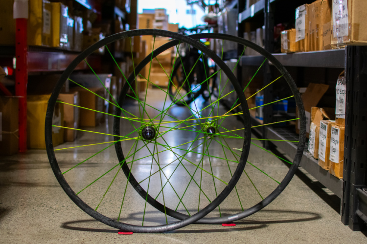 Two bicycle wheels with bright green spokes, leaning against each other in a store aisle filled with boxes. The background shows shelves stocked with various bike parts and accessories.