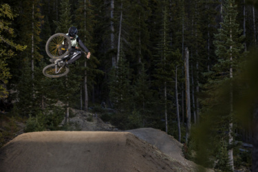 A mountain biker performs a mid-air trick above a dirt jump, surrounded by tall evergreen trees in a forested area. The rider is wearing protective gear and is captured against a natural backdrop. Trestle Bike Park mountain bike trail.