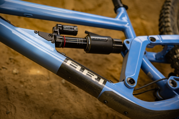 Close-up of a blue bicycle frame showing the suspension system and a sticker with the text "ARI." The background features a sandy surface, emphasizing the outdoor setting.