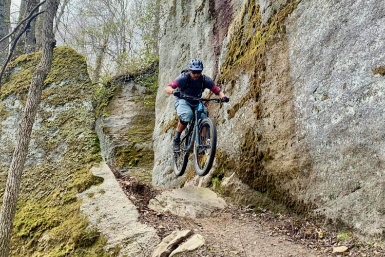A mountain biker performing a jump on a rocky trail, surrounded by moss-covered rocks and trees. The rider is wearing a helmet and protective gear, showcasing an action-packed moment in an outdoor forest setting.