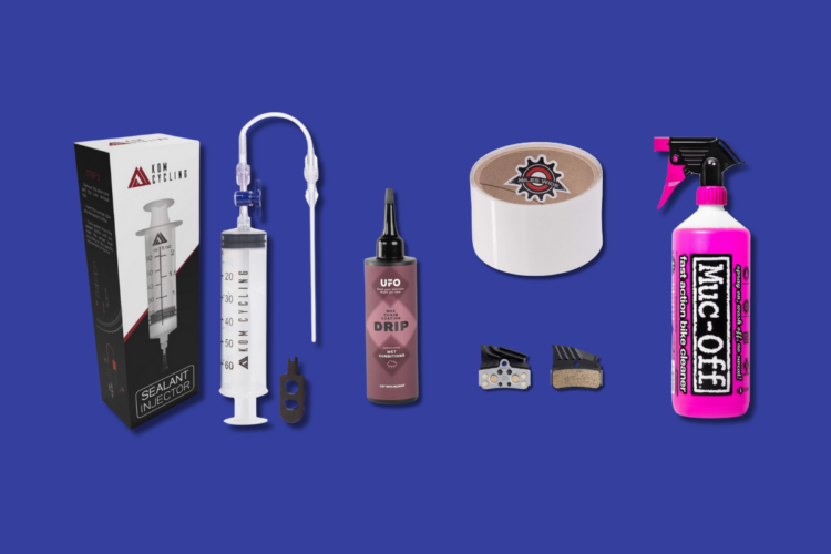 A selection of bike maintenance products on a blue background. The image features a sealant injector kit, a bottle of UFO bike drip lubricant for wet conditions, a roll of branded tape, a bottle of Muc-Off bike cleaner, and two types of brake pads.