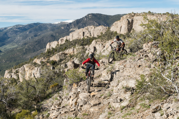 Two mountain bikers navigate a rocky trail surrounded by scenic mountains and greenery. One cyclist, wearing a red shirt and black pants, is leading the descent, while the other, dressed in a gray shirt and shorts, follows closely behind. The landscape features rugged terrain and distant snow-capped peaks under a clear blue sky. Cave Lake State Park mountain bike trail.