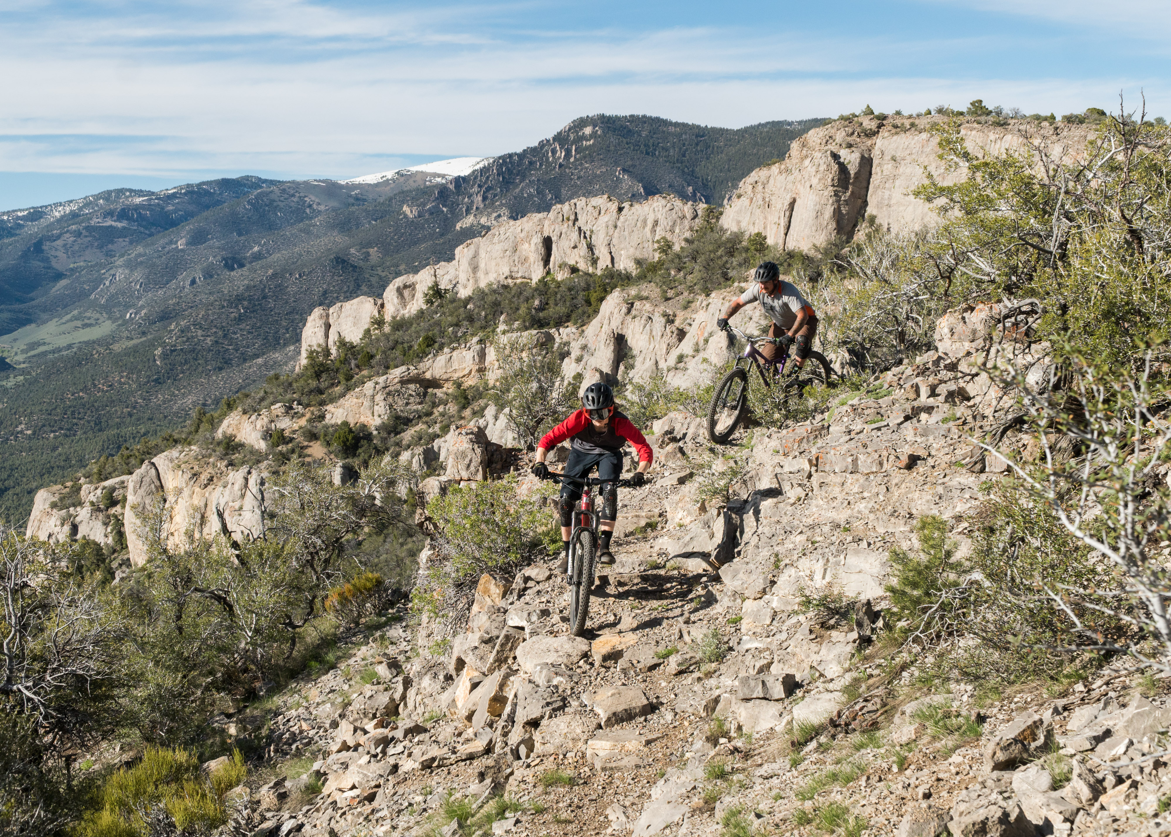 Two mountain bikers navigate a rocky trail surrounded by scenic mountains and greenery. One cyclist, wearing a red shirt and black pants, is leading the descent, while the other, dressed in a gray shirt and shorts, follows closely behind. The landscape features rugged terrain and distant snow-capped peaks under a clear blue sky. Cave Lake State Park mountain bike trail.