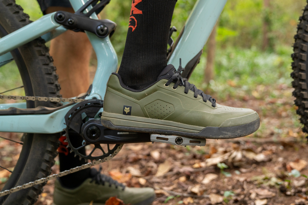 Close-up of a cyclist's foot wearing an olive green Fox Union flat mountain biking shoe, positioned on the pedal of a light blue mountain bike. The background features a natural, wooded setting with earthy tones and foliage.
