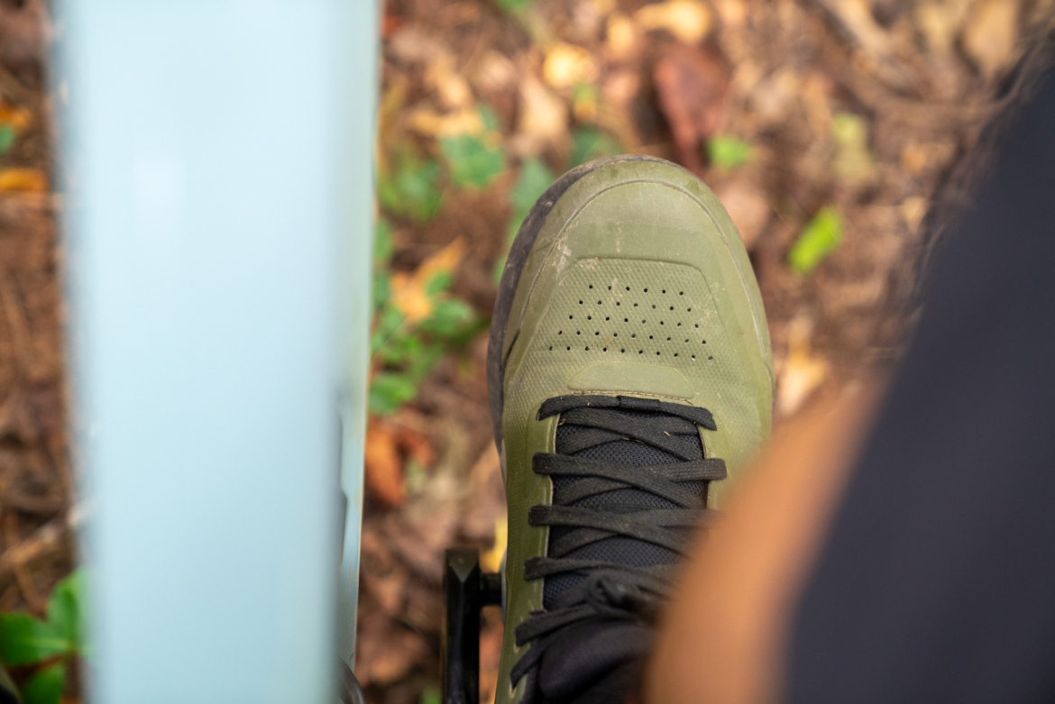 A close-up view of a green shoe resting on a pedal, with a blurred background of brown leaves and green foliage. The shoe features a textured surface and black laces.