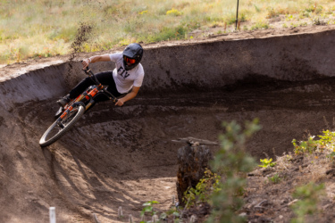 A mountain biker wearing a helmet and a white t-shirt leans into a dirt berm while riding an orange bike. Dust and particles are kicked up from the ground as he navigates the curve of the track, with a grassy landscape in the background. Bogus Basin mountain bike trail.