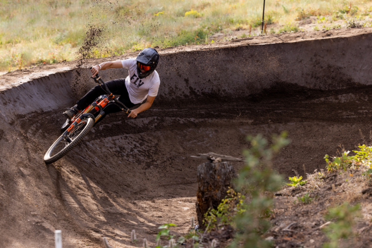 A cyclist in a helmet performs a sharp turn on a mountain bike track, kicking up dirt as they navigate a banked curve. The surrounding area features lush greenery and a sandy terrain with a stump visible in the foreground.