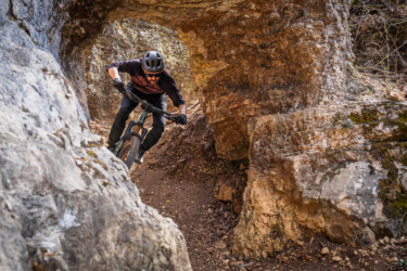 A mountain biker navigating through a natural stone archway on a dirt trail surrounded by rocky terrain and trees, showcasing dynamic movement and skill.