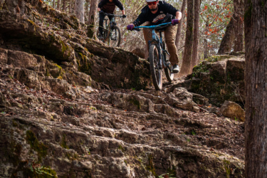 Two mountain bikers maneuver down a rocky trail in a wooded area, surrounded by trees with sparse leaves. The terrain is rugged, with large stones and patches of greenery. One cyclist is in the foreground, leaning forward as they navigate a steep section, while the other follows closely behind. The scene captures the excitement and challenge of mountain biking in nature. Great Passion Play Trails (GPPT) / Marble Flats mountain bike trail.