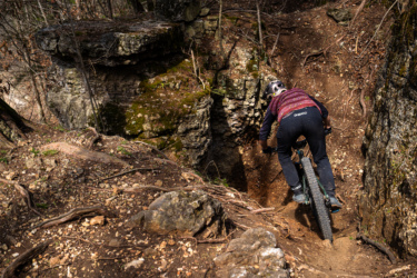 A mountain biker navigates a rocky trail in a wooded area, leaning forward as they descend a steep, narrow path surrounded by moss-covered rocks and tree roots. Great Passion Play Trails (GPPT) / Marble Flats mountain bike trail.