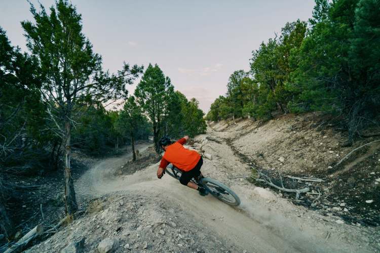 A mountain biker in an orange shirt leans into a turn on a dirt trail surrounded by trees, kicking up dust as he navigates a winding path through the rugged terrain.