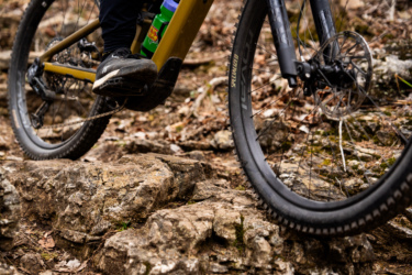Close-up image of a mountain bike navigating rocky terrain. The focus is on the bike's front wheel and the rider's foot positioned on the pedal, with a visible water bottle attached to the bike frame. The background features uneven rocks and scattered leaves, indicating an outdoor trail environment. Lake Leatherwood Gravity Project mountain bike trail.