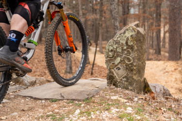 A close-up view of a mountain bike approaching a stone marker with the inscription "DH3." The image captures the bike's front wheel and part of the rider's leg, set against a wooded trail littered with fallen leaves. The background features a winding path leading deeper into the forest.