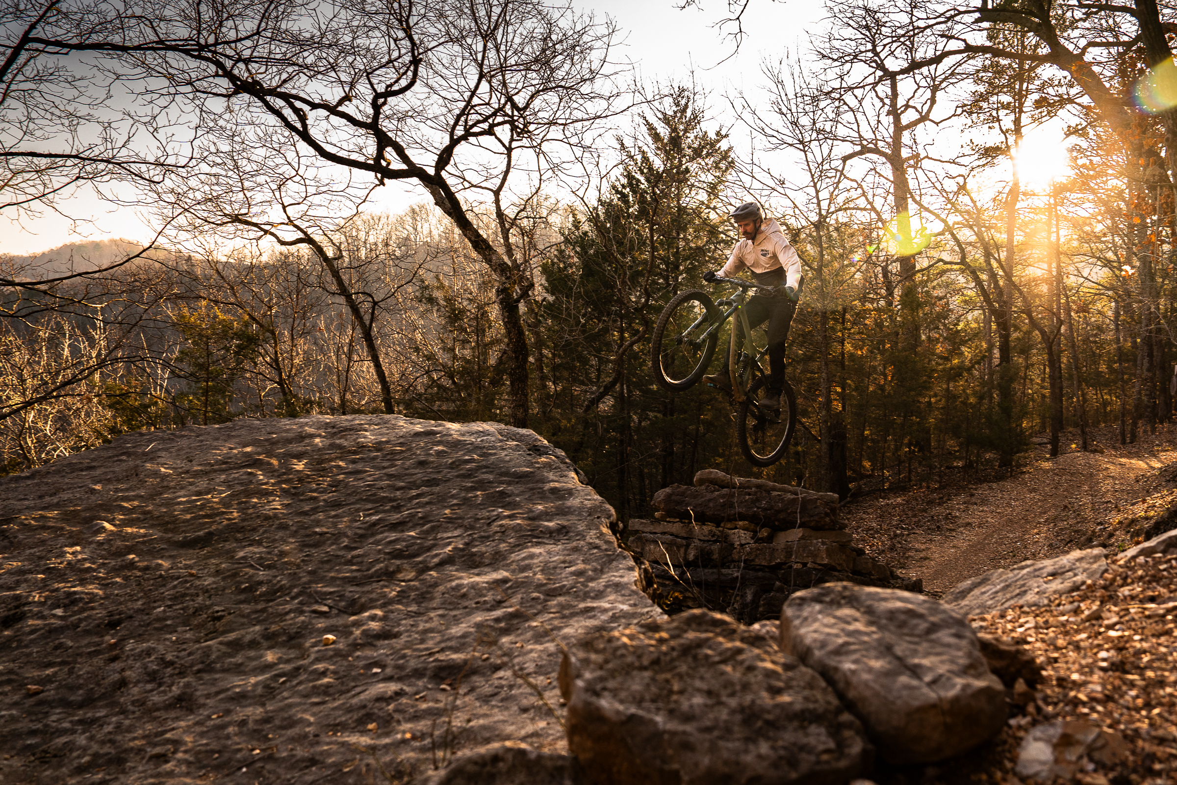 A mountain biker performing a jump off a rocky ledge in a wooded area during sunset. Trees are visible in the background, and warm sunlight filters through the branches, creating a dynamic and adventurous atmosphere. Lake Leatherwood Gravity Project mountain bike trail.