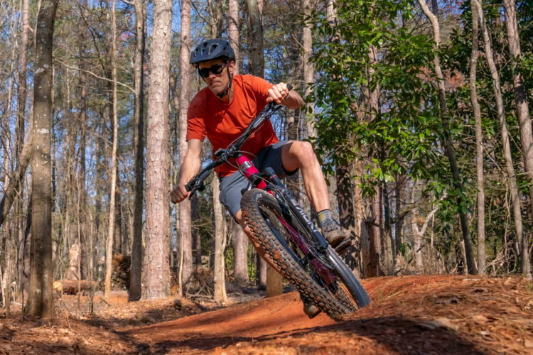 A mountain biker in an orange shirt and sunglasses navigates a dirt trail surrounded by trees, leaning into a turn on a mountain bike. The scene captures the action and thrill of outdoor cycling in a forested area.