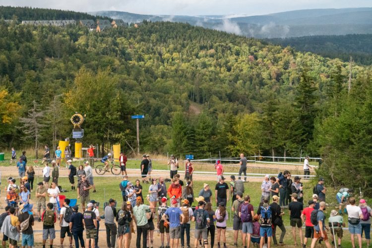A crowd of people gathered outdoors in Snowshoe, West Virginia, with a backdrop of rolling hills and trees. Some individuals are talking in groups, while others are engaged in activities like biking. Notable features include yellow structures and a signpost, with a mix of autumn colors in the foliage.