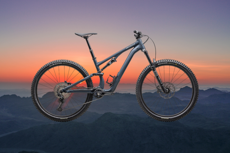 A mountain bike suspended in mid-air against a vibrant sunset backdrop, showcasing a mountainous landscape with layered hills and a gradient sky transitioning from deep purple to orange. The bike features thick tires and a sleek frame design, emphasizing its suitability for off-road terrain.