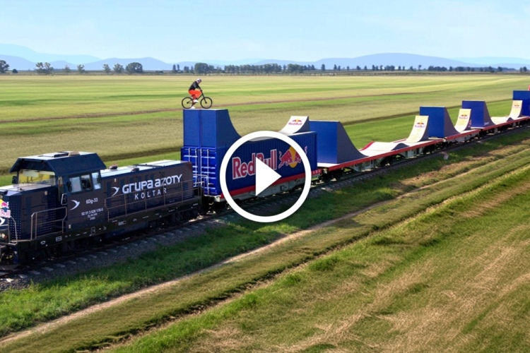 A train carrying large ramps for BMX biking, with a rider performing a stunt atop one of the ramps in a grassy, open landscape. The scene features a blue train locomotive with branding and a vibrant blue sky in the background.