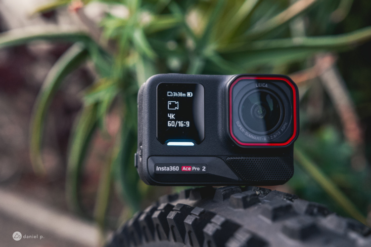 A compact Insta360 Ace Pro 2 camera positioned on a bicycle tire, featuring a digital display showing video settings including 4K resolution and 60 frames per second. The background consists of green foliage, providing a natural setting for the camera display.
