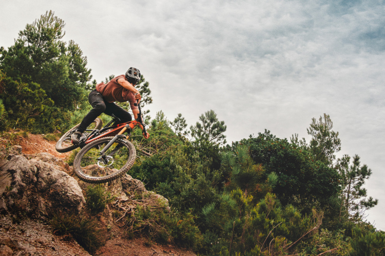 A mountain biker jumping over rocky terrain with trees in the background under a cloudy sky.