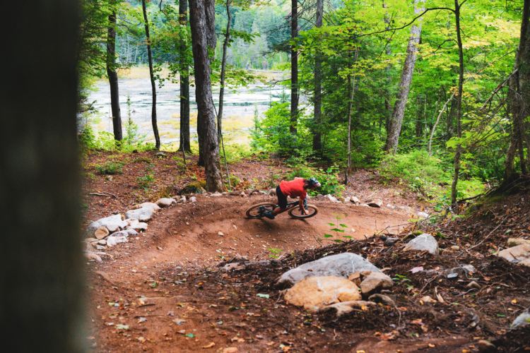 A mountain biker rides through a winding dirt trail in a lush forest setting, surrounded by trees and rocks. The cyclist, wearing a red shirt and a helmet, leans into a turn as the bike navigates the terrain, with a blurred background of greenery and a glimpse of a water body.