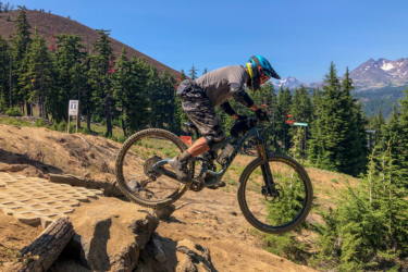 A mountain biker in a blue helmet and gray shirt jumps off a rocky ledge on a dirt trail surrounded by lush green trees and mountains in the background, under a clear blue sky. Mount Bachelor Bike Park mountain bike trail.