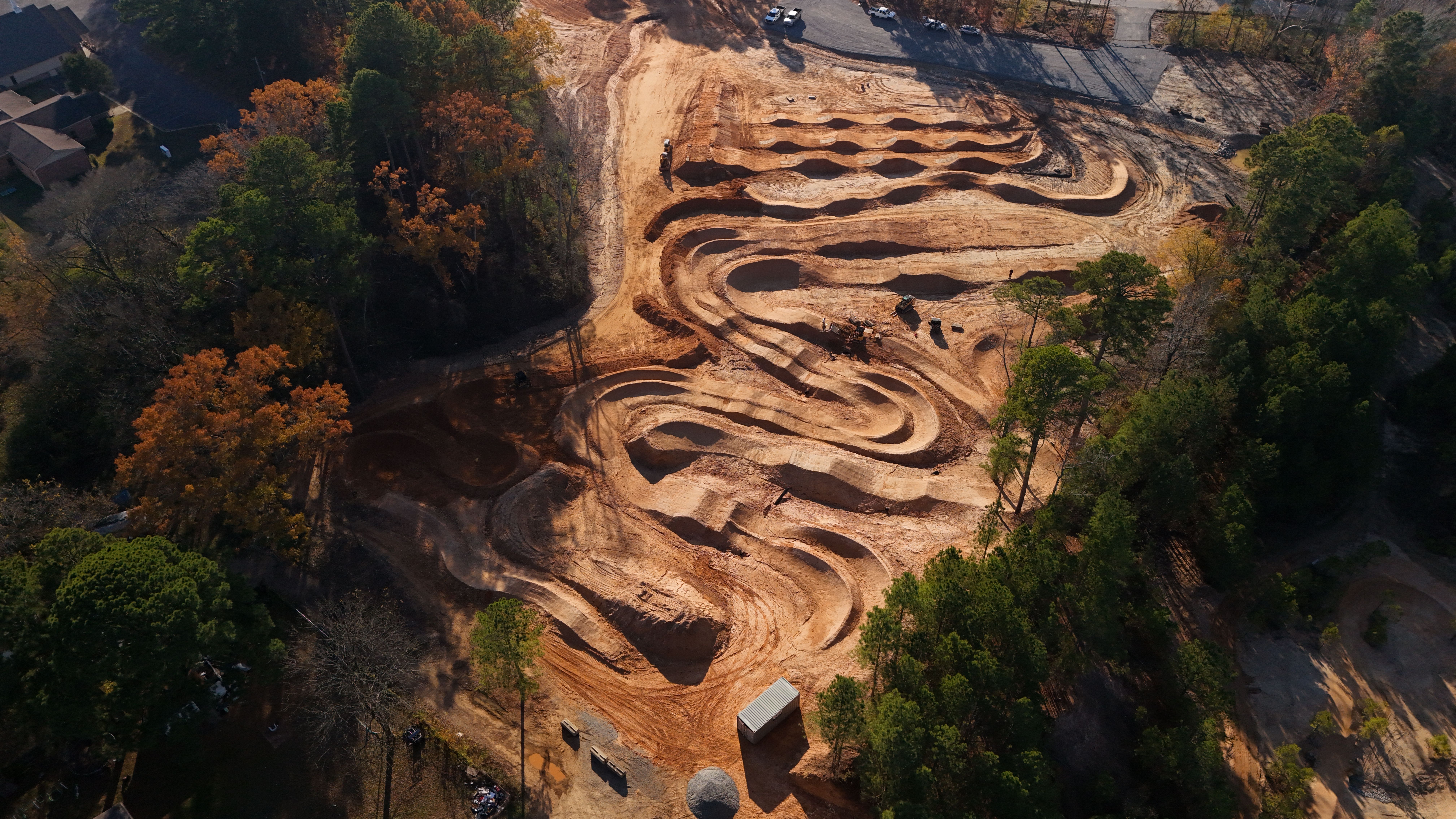 Aerial view of a recently constructed dirt bike track surrounded by trees, featuring winding paths and jumps. The landscape shows a mix of orange and brown earth with some greenery, highlighting the layout of the track and nearby vehicles. Benton Bike Park mountain bike trail.