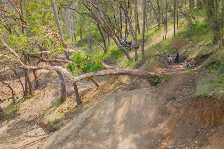 A mountain biker performing a trick in mid-air above a dirt jumping ramp surrounded by trees, with two bystanders watching nearby. The scene captures the excitement of outdoor biking in a natural setting.