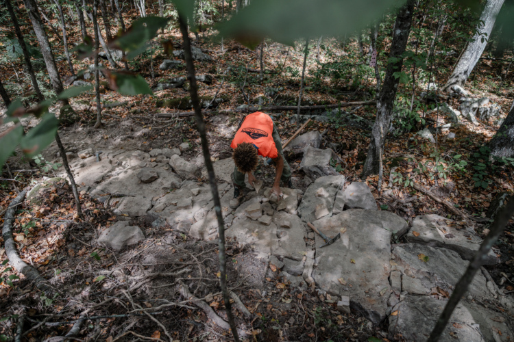 A person wearing an orange shirt is crouched down on a rocky path in a forested area, examining stones and rocks among fallen leaves and tree branches. The scene is surrounded by trees, showcasing a natural environment.