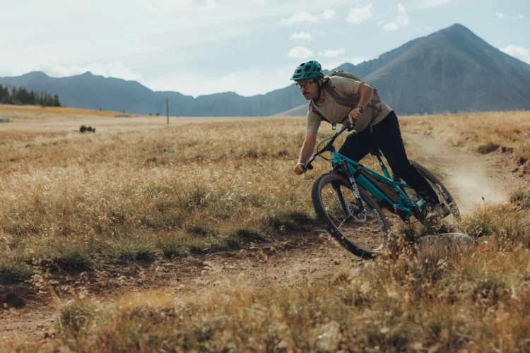 Joey Schusler riding a turquoise mountain bike with Enve Series M wheels on a dirt trail in a grassy open area, with mountains in the background and a partly cloudy sky. The rider is leaning into a turn, kicking up dust from the trail.