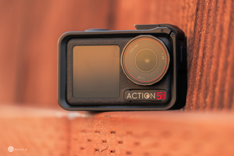 A close-up image of a black action camera resting on a wooden surface, displaying its lens and control buttons. The camera is labeled "ACTION 5 PRO." The background features a warm, blurred wooden texture.
