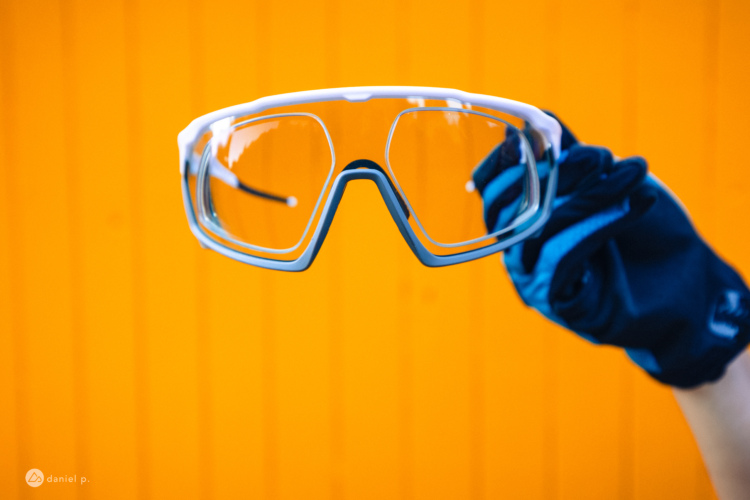 A pair of clear sports glasses is being held up against a bright orange background. The glasses have a modern design with a wide frame and are being grasped by a hand wearing a blue glove.