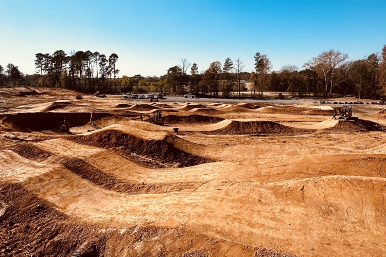 Construction site of a dirt bike or BMX track, featuring a series of dirt mounds, jumps, and berms, under a clear blue sky with trees in the background. Several construction vehicles are visible in the distance.