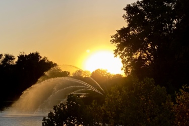 Sunset over a calm lake, with a fountain spraying water, surrounded by silhouettes of trees. The sky is filled with warm hues of orange and yellow as the sun sets on the horizon. Western University trails mountain bike trail.