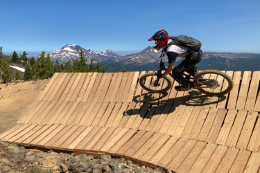A mountain biker riding on a wooden ramp, skillfully maneuvering through a downhill course with picturesque mountain ranges in the background. The scene features a clear blue sky and lush trees surrounding the area, showcasing a vibrant outdoor setting for mountain biking. Mount Bachelor Bike Park mountain bike trail.
