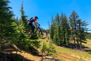 A mountain biker jumps over a log in a forested area, surrounded by tall trees against a clear blue sky. The cyclist is in mid-air, showcasing an adventurous moment in nature. Mount Bachelor Bike Park mountain bike trail.