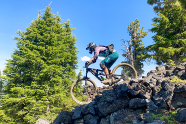 A mountain biker navigating rocky terrain on a sunny day, surrounded by tall green trees and a clear blue sky. The biker is wearing a helmet and protective gear, showcasing an active outdoor adventure. Mount Bachelor Bike Park mountain bike trail.