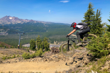 A mountain biker performing a jump on a downhill trail surrounded by pine trees, with a scenic mountain backdrop and blue sky. A ski lift can be seen in the distance amidst the lush green landscape. Mount Bachelor Bike Park mountain bike trail.