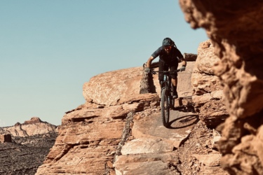 A mountain biker navigating rocky terrain against a clear blue sky. The cyclist is leaning forward on a rugged path surrounded by large boulders and desert landscape, showcasing an adventurous outdoor scene. Hawks Glide mountain bike trail.