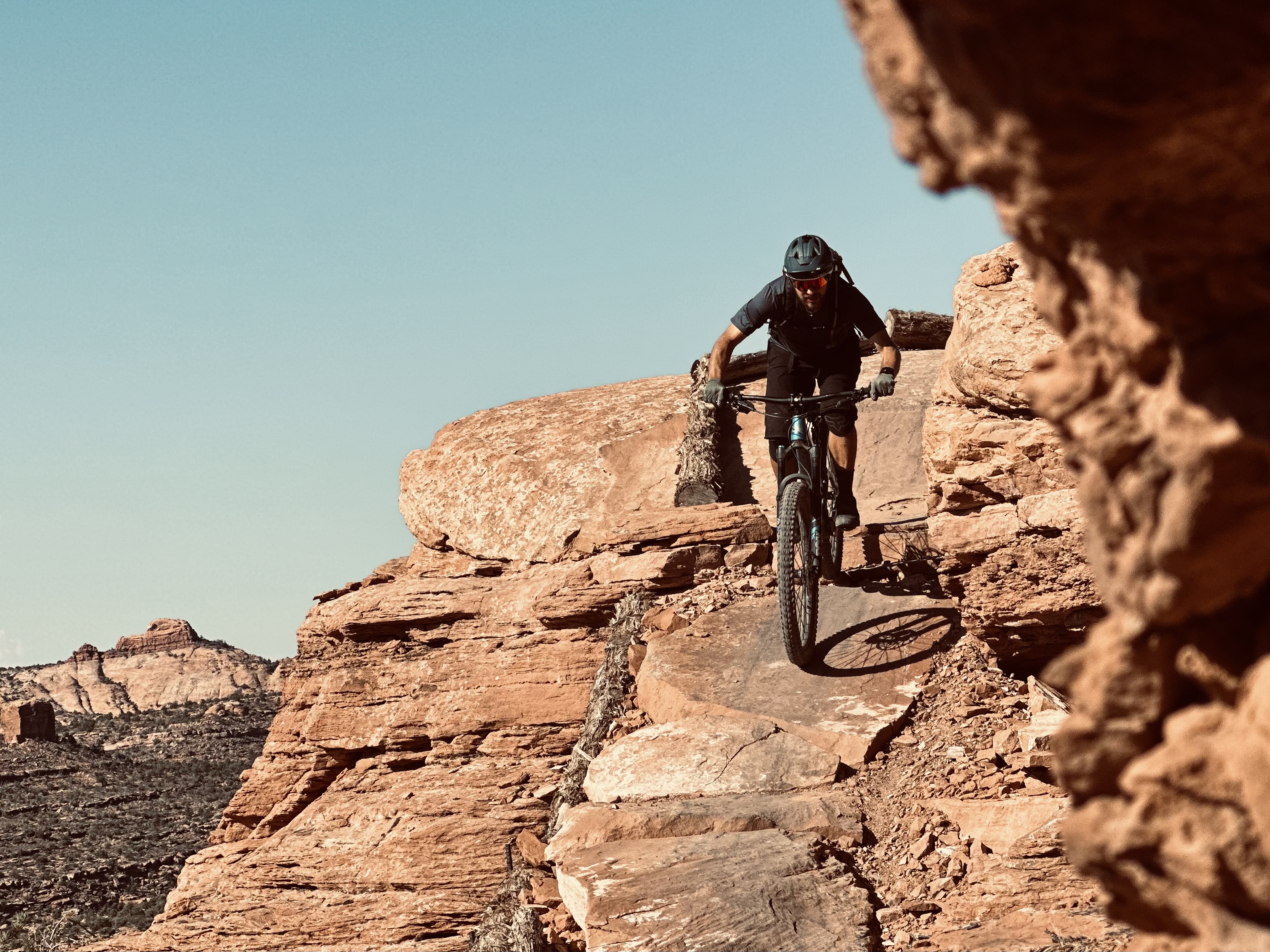 A mountain biker navigating rocky terrain against a clear blue sky. The cyclist is leaning forward on a rugged path surrounded by large boulders and desert landscape, showcasing an adventurous outdoor scene. Hawks Glide mountain bike trail.